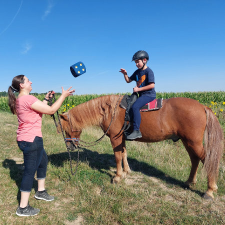 Ein Junge auf einem Pferd und eine Frau werfen sich einen großen Würfel zu (Reittherapie).